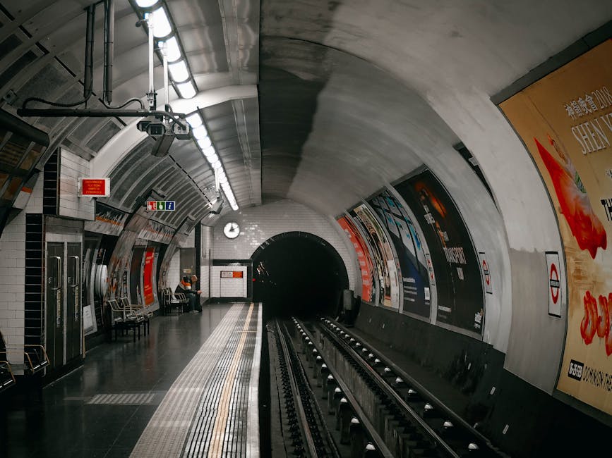 An empty underground station corridor with curved tiled walls and a tiled floor, illuminated by overhead fluorescent lights. The walls are cream-colored with a grid pattern, and the ceiling features s