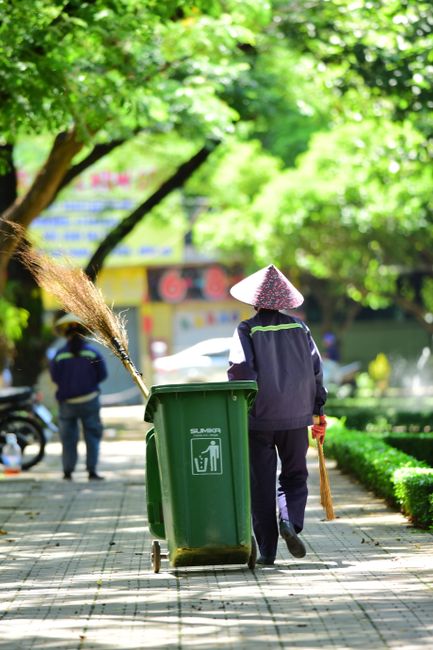 A waste management worker wearing a high-visibility yellow jacket with the words 'Waste Management Services' and 'Working Safely for Dublin City' printed on the back, is collecting trash from a green 